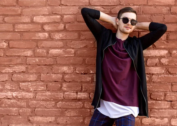 Хлопець моди в окулярах стоїть біля цегляної стіни з потрісканою фарбою — стокове фото