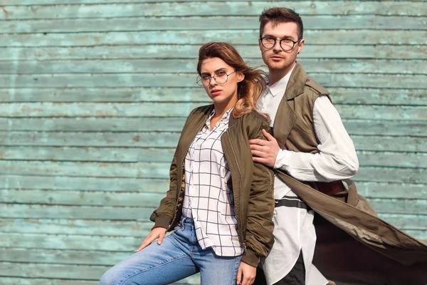 Пара моди в окулярах в зеленому одязі позує на синій дерев'яній стіні — стокове фото