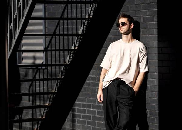 Fashion guy stands posing near a black brick wall in sunglasses — Stock Photo, Image