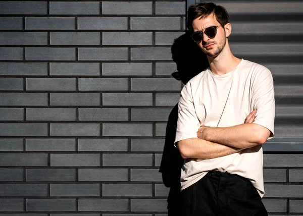 Hombre de moda se para posando cerca de una pared de ladrillo negro en gafas de sol —  Fotos de Stock