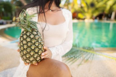 Kız, tropik iklimdeki havuzun arka planında ananas tutuyor.