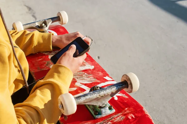 Close-up de um adolescente vestido com uma calça de camisola e tênis sentado em um parque de skate segurando um telefone e um skate — Fotografia de Stock