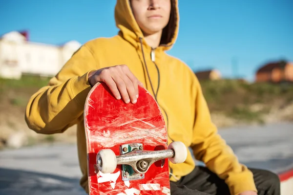 Close-up of a teenager dressed in a jeans hoodie sitting in a skate park and holding a skateboard — Stock Photo, Image