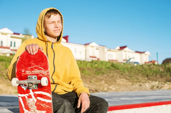 Close-up de um adolescente vestido com capuz jeans sentado em um parque de skate e segurando um skate — Fotografia de Stock