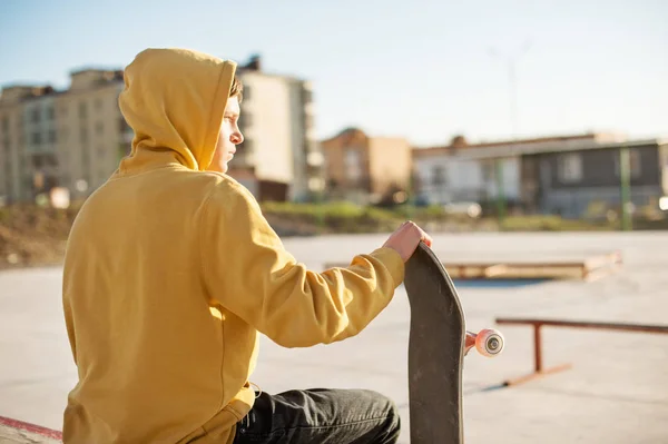 Close-up de um adolescente vestido com capuz jeans sentado em um parque de skate e segurando um skate — Fotografia de Stock