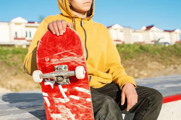 Close-up de um adolescente vestido com capuz jeans sentado em um parque de skate e segurando um skate — Fotografia de Stock