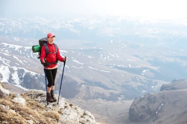 Kayalar ve uzak diyarlardan fon karşı dağlarda şapkalı bir sırt çantası ve dinlenmek için katlanmış bir halı ile Kuzey yürüyüşü için sopa ile ince bir kız duruyor