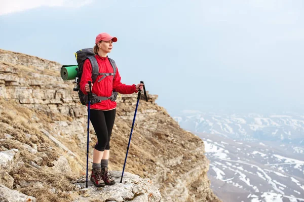 Štíhlá dívka v čepici s holemi pro Nordic walking s batohem a skládané koberec pro relaxaci stojí v horách na pozadí skal a dalekých zemí — Stock fotografie