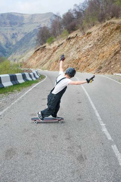 Longboarder a longboard, overall, sisak és kesztyű végez egy stand-up dia sebességgel, míg úton egy hegyi szerpentin a hegyekben, egy gyönyörű hegyi táj a háttérben — Stock Fotó