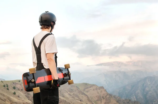 Un joven en un casco con guantes con una tabla en las manos y vestido con un combo está parado en un precipicio en lo alto de las montañas contra el telón de fondo de la puesta de sol — Foto de Stock