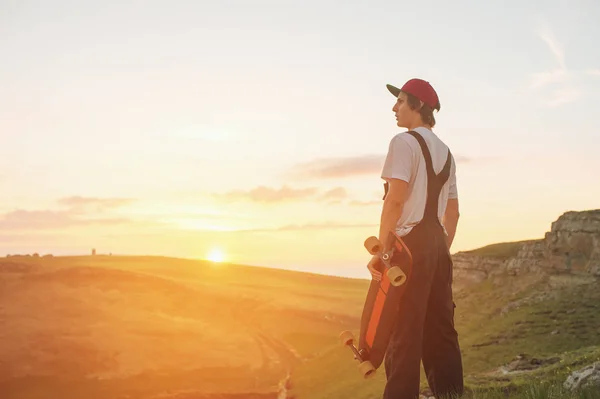 Un tipo con una gorra y un kabenison con una tabla larga en sus manos está de pie en la naturaleza contra la puesta de sol — Foto de Stock
