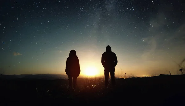 Ett par personer man och kvinna står vid solnedgången av månen under stjärnklar himmel med ljusa stjärnor och en Vintergatan. Siluett foto mot den stjärnklara natthimlen — Stockfoto