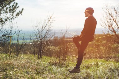 Bir adam gözlüklü bir şapka ve spor ayakkabı slackline üzerinde oturan yaşta denge yakalar ve hayat parkta niteliğine sahiptir