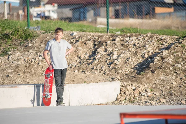 Ein Teenager steht in einem Skatepark in einem grauen T-Shirt mit einem Skateboard in der Hand vor der Kulisse des Stadtrands — Stockfoto