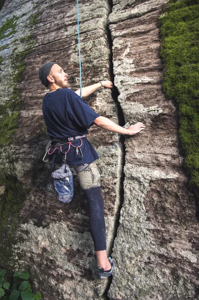 A young hipster is engaged in rock climbing with insurance on rocks with green moss — Stock Photo, Image