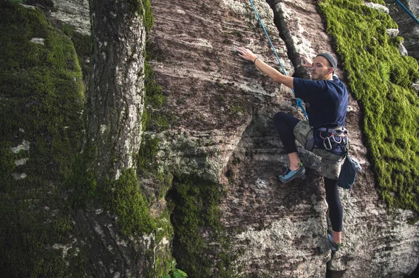 Молодой хипстер занимается скалолазанием со страховкой на скалах с зеленым мхом — стоковое фото