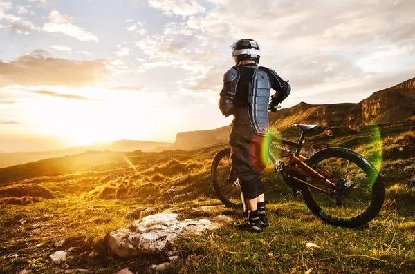 El piloto en equipo de protección completa en la bicicleta mtb está montando hacia el atardecer en los rayos del sol puesta del sol contra el fondo de las rocas del sol poniente y las nubes —  Fotos de Stock