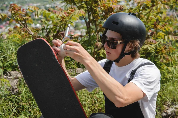 Um jovem de óculos de sol e macacão com um capacete na cabeça muda suas rodas em seu longboard sob o céu aberto — Fotografia de Stock