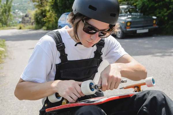 A young man in sunglasses and overalls with a helmet on his head changes his wheels on his longboard under the open sky — Stock Photo, Image