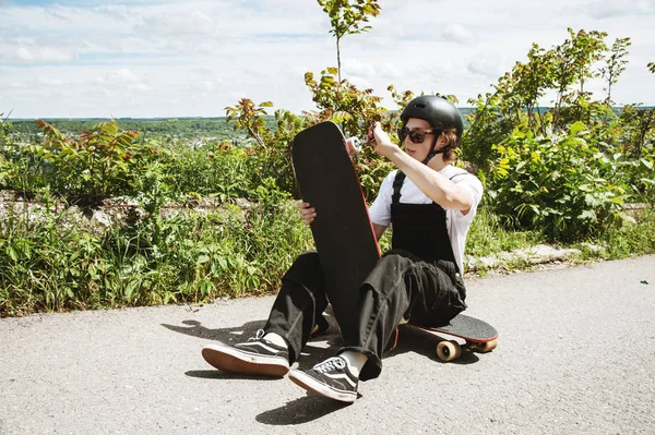 Um jovem de óculos de sol e macacão com um capacete na cabeça muda suas rodas em seu longboard sob o céu aberto — Fotografia de Stock