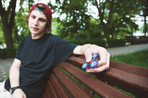 A long-haired hipster in a cap sits on a bench and spins a fidget-spinner — Stock Photo, Image