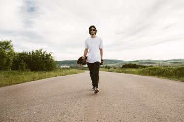 Bir longboard kask ve güneş gözlüğü onun elinde olan bir adam bir asfalt banliyö yolda yürüyor