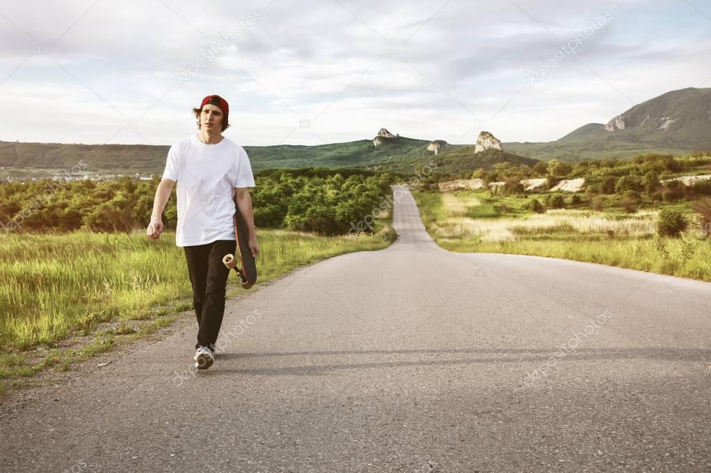 A man in a cap and with a long board in his hands is walking on a country road