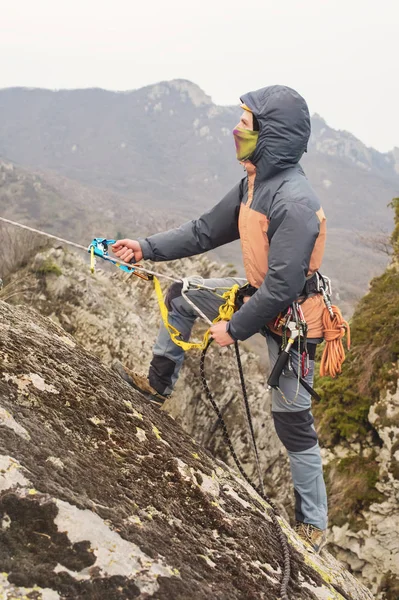 Der junge Mann erklimmt den Felsen an einem Seil mit Sicherheitsgurten, Versicherung und Seil, in voller Bergsteigerausrüstung und Helm — Stockfoto