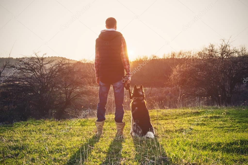 A man with a beard walking his dog in the nature, standing with a backlight at the rising sun, casting a warm glow and long shadows against the background of the gorge and trees.