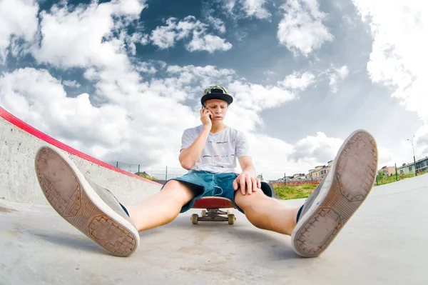 Junger Skater sitzt auf Skateboard im Park und telefoniert — Stockfoto