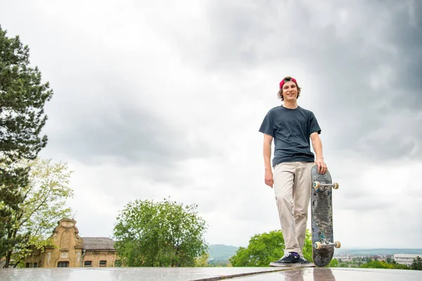 Ein junger Skater steht mit einem Skateboard vor dem Hintergrund der Landschaft der Stadt — Stockfoto