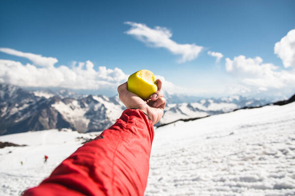 A mans hand holds an apple with a bite on the background of snow-covered mountains and snow underfoot.