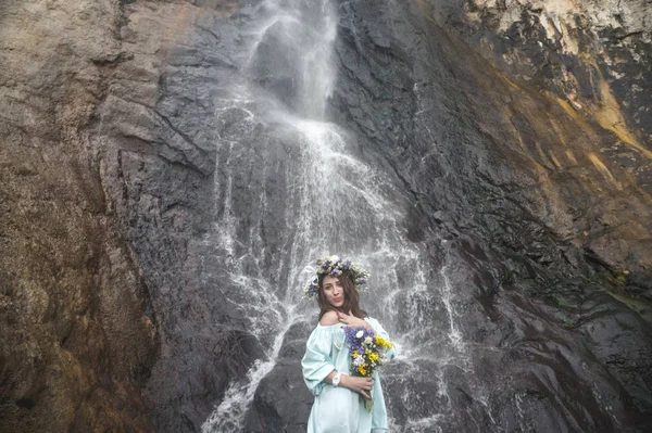 Retrato de uma menina com uma coroa de flores na cabeça e um buquê de flores nas mãos contra uma cachoeira — Fotografia de Stock