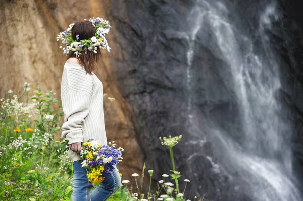 Retrato de uma menina com uma coroa de flores na cabeça e um buquê de flores nas mãos contra uma cachoeira — Fotografia de Stock