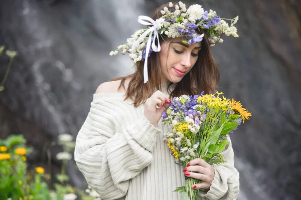 Retrato de uma menina com uma coroa de flores na cabeça e um buquê de flores nas mãos contra uma cachoeira — Fotografia de Stock