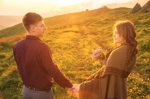 Una pareja joven. El tipo lleva a una chica rizada envuelta en un cuadros con un ramo de flores al atardecer. . — Foto de Stock