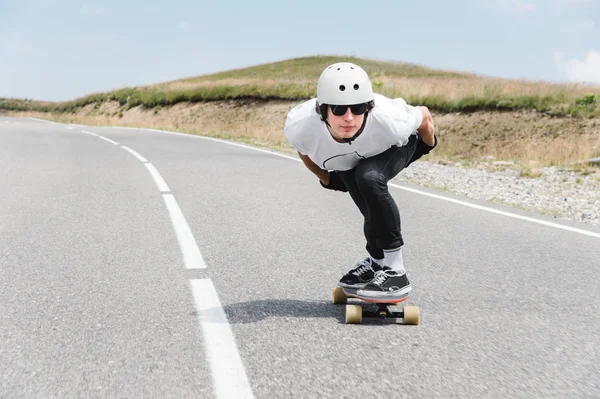 Egy srác visel sisakot és napszemüveg a lovaglás a longboard úton — Stock Fotó