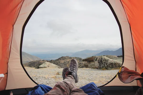 Legs of a man resting in boots for mountain tracking in travel tent against the backdrop of mountains and valleys with noisy clouds
