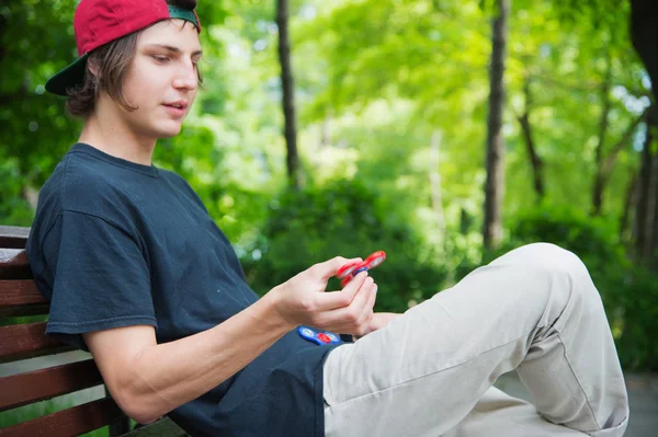Un hipster de pelo largo con una gorra se sienta en un banco y hace girar a un agitador — Foto de Stock