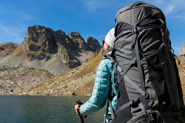 Una ragazza escursionista in occhiali da sole e un cappello con zaino e attrezzatura da montagna con trekking di monitoraggio nelle sue mani guarda la splendida vista di un lago di alta montagna in un arcipelago nel Caucaso settentrionale — Foto Stock
