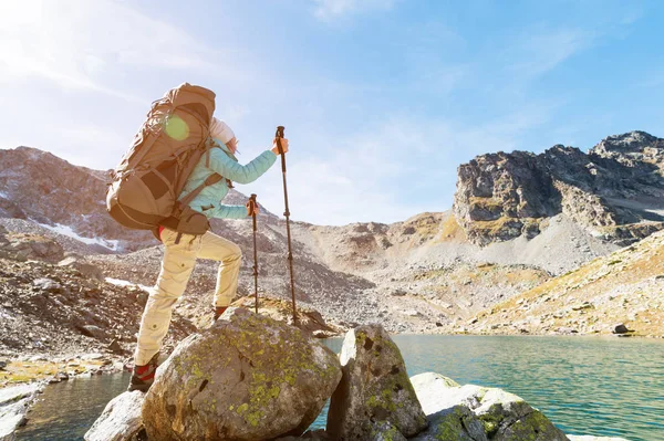 Una ragazza escursionista in occhiali da sole con uno zaino e bastoni di monitoraggio sale a una roccia alta sullo sfondo di rocce e un lago di alta montagna — Foto Stock