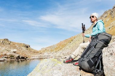 Ceket ve şapka ile bir sırt çantası ve dağ ekipman için izleme içinde elleri kolları ile aşağı güneş gözlüğü takıyor bir turist kız yüksek dağ Gölü üzerinde oturuyor