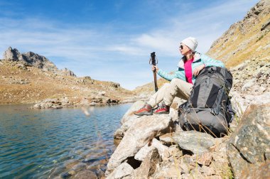 Ceket ve şapka ile bir sırt çantası ve dağ ekipman için izleme içinde elleri kolları ile aşağı güneş gözlüğü takıyor bir turist kız yüksek dağ Gölü üzerinde oturuyor