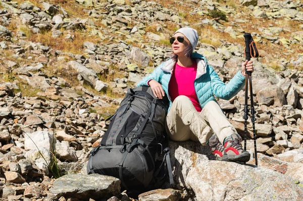 Turistické dívka, která nosí sluneční brýle bundy a čepice s batohem a horských zařízení s úchyty pro sledování v jejích rukou se sedí vysoko v horách — Stock fotografie