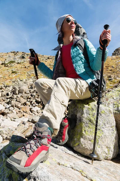 Turistické dívka, která nosí sluneční brýle bundy a čepice s batohem a horských zařízení s úchyty pro sledování v jejích rukou se sedí vysoko v horách — Stock fotografie