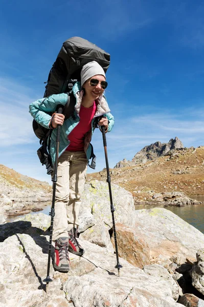 Tramp dívka v sluneční brýle s batohem a sledování hole stoupne na vysoké skále na pozadí skal a vysoké horské jezero — Stock fotografie