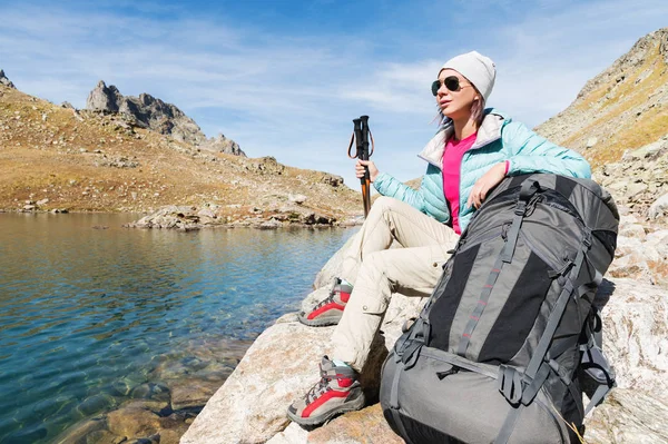Turistické dívka, která nosí sluneční brýle bundy a čepice s batohem a horských zařízení s úchyty pro sledování v jejích rukou se sedí na vysoké horské jezero — Stock fotografie