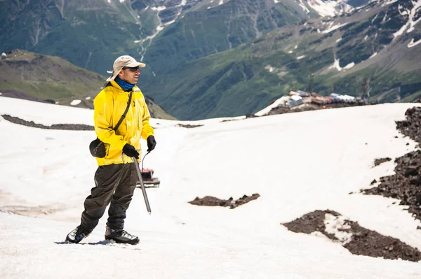 Um montanhista em um boné com óculos e um picador de gelo fica em um fundo de uma montanha coberta de neve — Fotografia de Stock