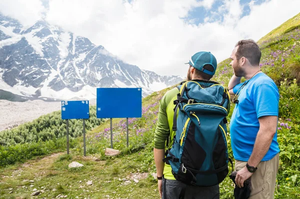 Mock up. Two tourists with backpacks stand in the mountains and look at the empty board.