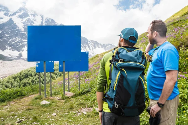 Mock up. Two tourists with backpacks stand in the mountains and look at the empty board.